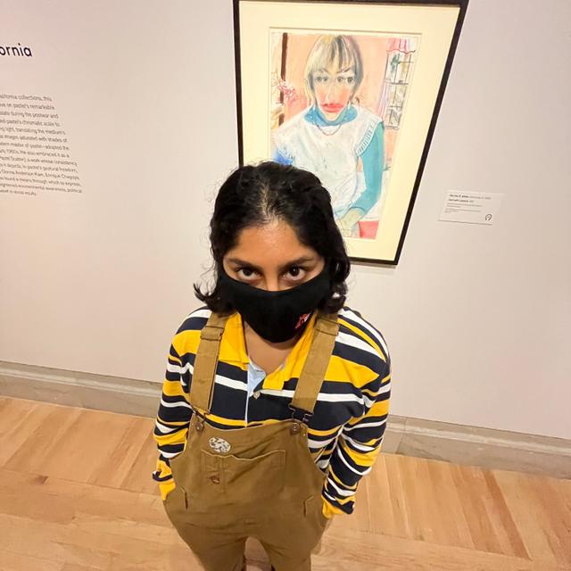 maya in overalls standing in front of an art piece with a threatening expression